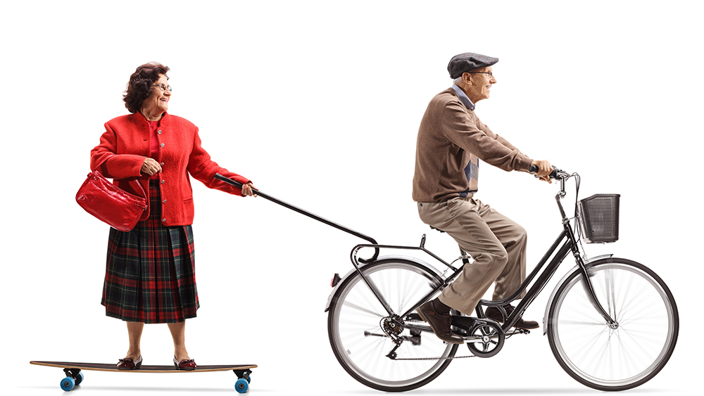Kuvituskuva, jossa iäkkäämpi iloinen nainen roikkuu kävelykepin avulla skeittilaudallaan iloisen iäkkään miehen polkupyörän tavaratelineessä ajon aikana.