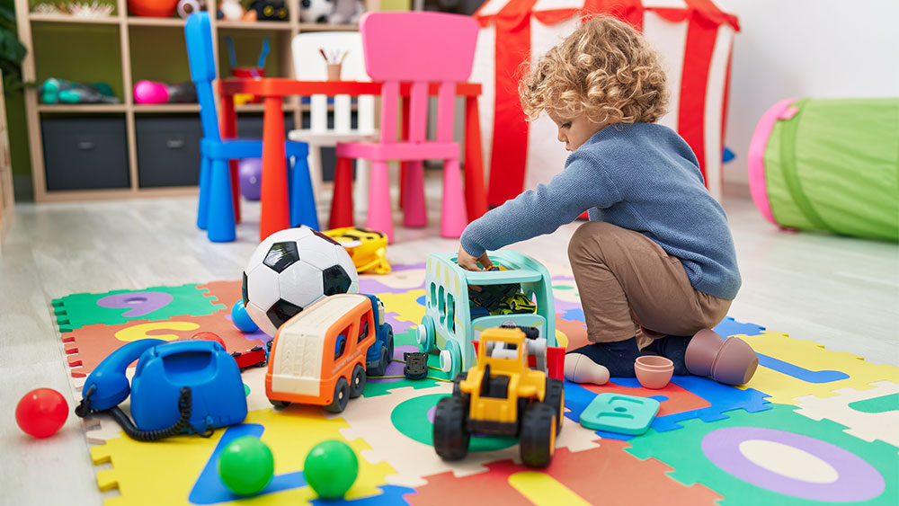 Kuvassa kiharatukkainen taaperoikäinen lapsi leikkii lattialla liikennematon päällä useita erilaisia leluja ympärillään.