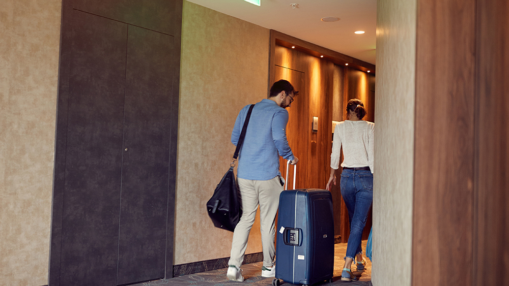 Nainen ja mies selät kameraan päin kävelemässä hotellihuoneessa matkalaukkujen kanssa