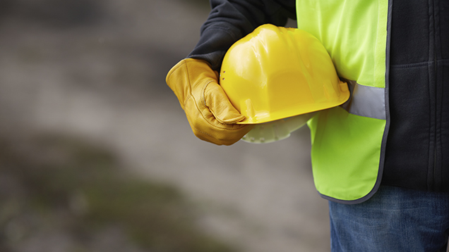 Kuvituskuva: Rakennustyömies pitelee keltaista kypärää kainalossaan hanskakäsin.