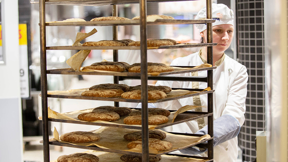 Kuvassa naisoletettu leipomotyöntekijä työntää ruisreikäleipiä sisältävää metallirullakkoa uunilta pakkaamoon.