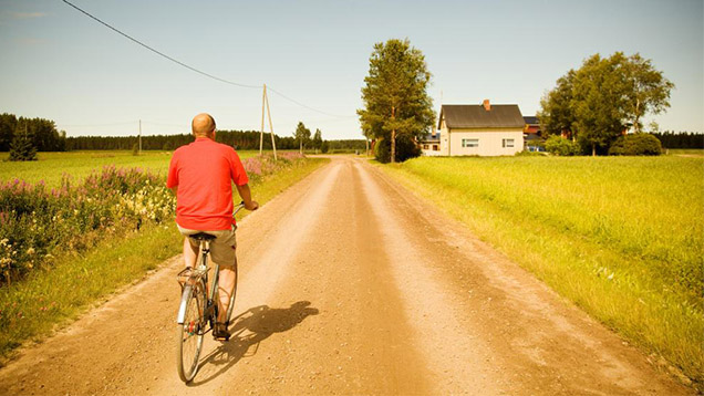 Yksinäinen ihminen ajaa polkupyörällä peltojen keskellä hiljaisella kylätiellä.
