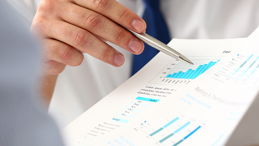 Ihmisen kädet, joissa kynä ja taustalla papereita, joissa erilaisia kaavioita ja tekstiä.