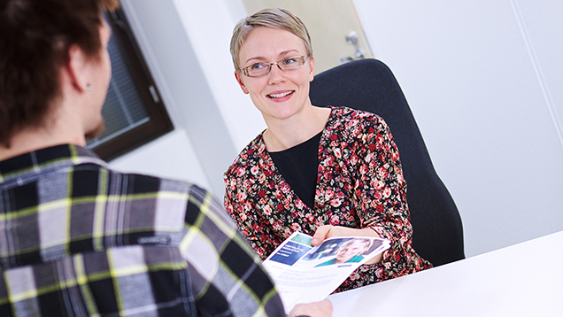 The image shows a customer service situation in which a woman is handing a leaflet to a young man.