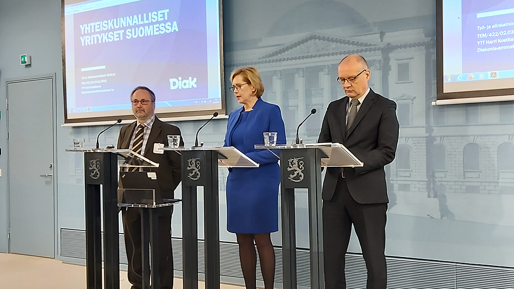 Tutkija Harri Kostilainen, työministeri Tuula Haatainen ja ohjausryhmän puheenjohtaja Kimmo Ruth seisovat tiedotustilaisuudessa mikrofonien äärellä.